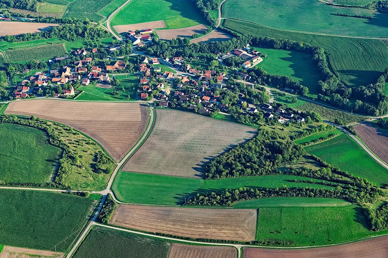 Luftaufnahme von Kirnberg im Sommer: rote Dächer zwischen Bäumen, umgeben von Feldern, Wiesen, Hecken und Waldstreifen am Fuß der Frankenhöhe